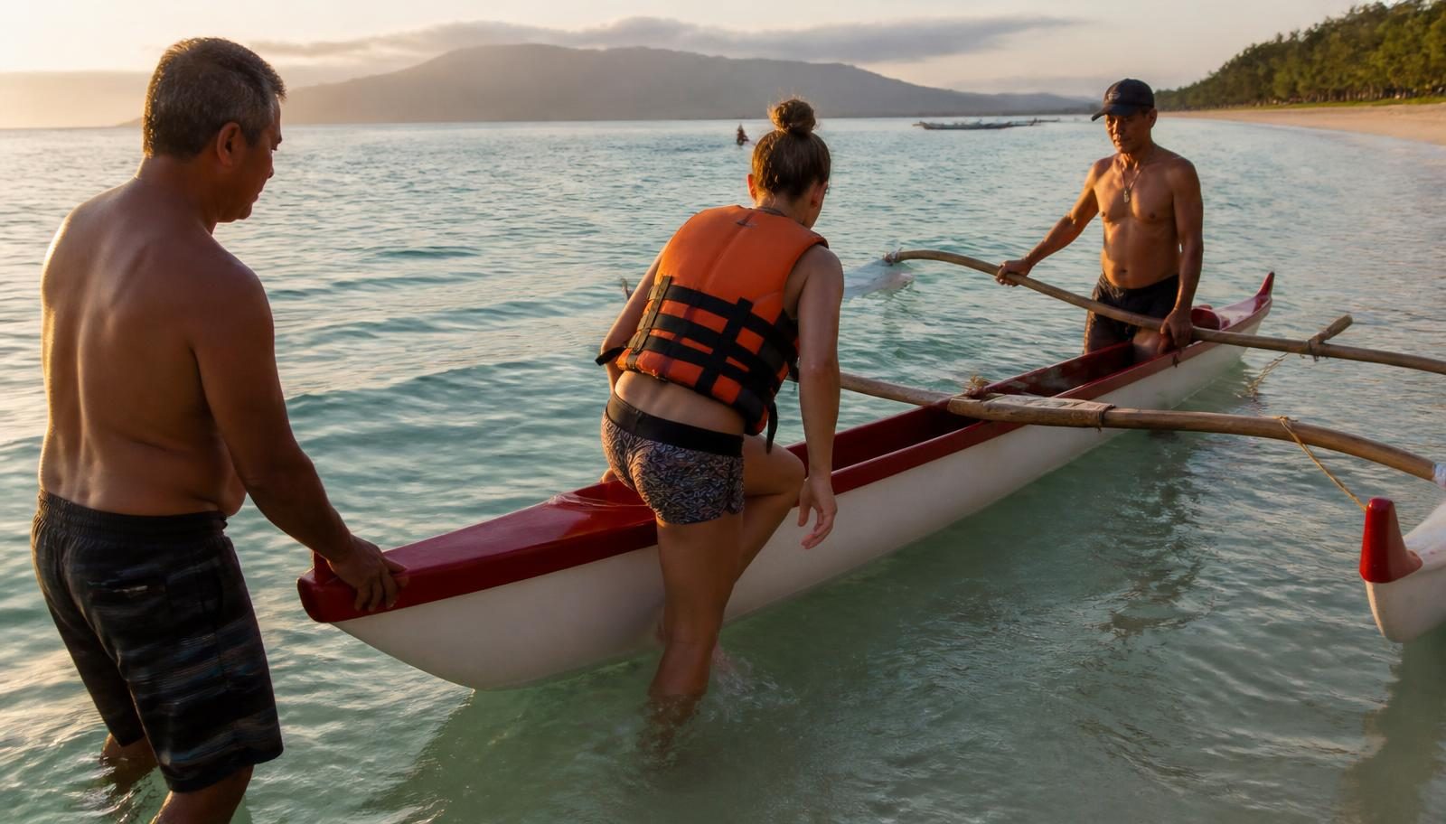 Visitante de colete salva-vidas entrando em uma canoa havaiana segurada por dois remadores experientes ao amanhecer